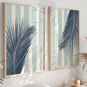 Op de afbeelding: Twee ingelijste wandkunstwerken met licht houten frames. Elk heeft een marineblauw palmbladontwerp over een gestreepte achtergrond van lichtblauw en crème. Het kunstwerk wordt tentoongesteld boven een houten bank met decoratieve items.