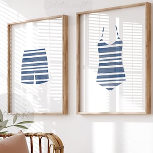 May include: Two framed prints featuring watercolor illustrations of swimwear. One print shows blue and white striped swim trunks, and the other depicts a blue and white striped swimsuit. The frames are made of light brown wood.