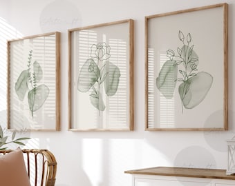 Puede incluir: Tres impresiones enmarcadas con dibujos minimalistas de plantas verdes sobre un fondo blanco. Las impresiones están colgadas en una pared blanca sobre una cómoda blanca con dos cajones. Una silla de mimbre con un cojín marrón y una manta blanca está en primer plano.