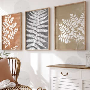 May include: Three framed botanical prints with fern and leaf designs in neutral tones of brown, gray, and beige. The prints hang above a wicker chair with a terracotta pillow and a white blanket. A white cabinet and a woven basket are also in the room.