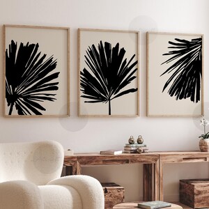 Może przedstawiać: Trzy oprawione wydruki artystyczne z czarnymi abstrakcyjnymi wzorami liści palmowych na beżowym tle. Wydruki są w drewnianych ramach, tworząc minimalistyczną i nowoczesną estetykę. Grafika jest eksponowana nad drewnianą konsolą.