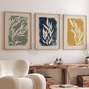 Puede incluir: Tres impresiones enmarcadas con siluetas blancas de ramas frondosas sobre un fondo de color. Las impresiones están en un salón con un sillón blanco, una mesa de café de madera y una consola de madera.