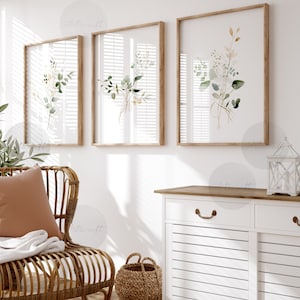 Puede incluir: Un armario blanco con dos cajones y dos puertas, con una tapa de madera y tiradores marrones. Una cesta de mimbre se encuentra en el suelo delante del armario. Una silla de mimbre marrón con un cojín beige y una manta blanca se encuentra en primer plano. Tres impresiones de acuarela enmarcadas de hojas verdes y detalles dorados cuelgan en la pared detrás del armario.