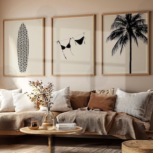 May include: Three framed prints on a beige wall above a couch with brown throw pillows. The prints feature a black and white silhouette of a surfboard, a palm tree, and a bikini hanging on a clothesline.