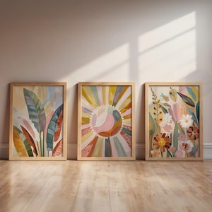 Op de afbeelding: Drie ingelijste kunstdrukken met botanische en abstracte ontwerpen. Het kunstwerk bevat kleurrijke afbeeldingen van bladeren, een zonnestraal en verschillende bloemen. Elke print zit in een lichtgekleurde houten lijst, leunend tegen een muur op een houten vloer.