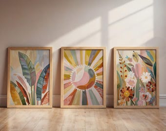 May include: Three framed art prints with botanical and abstract designs. The artwork includes colourful depictions of leaves, a sunburst, and various flowers. Each print is in a light-coloured wooden frame, leaning against a wall on a wooden floor.