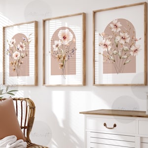 May include: Three framed floral art prints. Each print showcases a bouquet of watercolour flowers in shades of pink, cream, and beige, set against a soft pink arch background. The frames are made of light-coloured wood, creating a cohesive set.