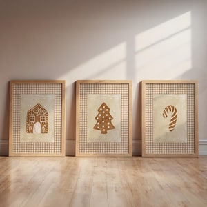 May include: Three framed prints featuring Christmas-themed designs. The prints showcase a gingerbread house, a Christmas tree, and a candy cane, all in a warm brown hue against a checkered background. The frames are made of light-colored wood.