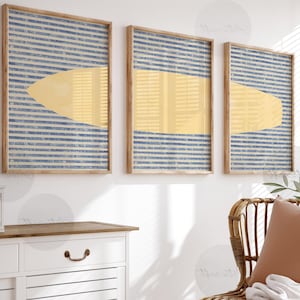 Op de afbeelding: Drie ingelijste abstracte kunstprints. Elke print heeft een gele vorm over een blauw-wit gestreepte achtergrond. De frames zijn gemaakt van lichtgekleurd hout. De kunst wordt op een witte muur getoond.