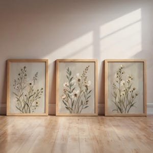 Peut inclure: Trois impressions d'art botanique encadrées. Chaque impression présente une illustration à l'aquarelle de fleurs blanches délicates et de feuillage vert sur un fond neutre. Les cadres sont en bois clair, exposés sur un sol en bois.