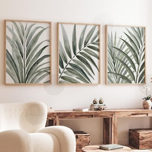 Op de afbeelding: Drie ingelijste prints met aquarel illustraties van groene palmbladeren. Het kunstwerk hangt aan een witte muur boven een houten console en een crèmekleurige fauteuil, wat een serene, natuurlijke esthetiek creëert.