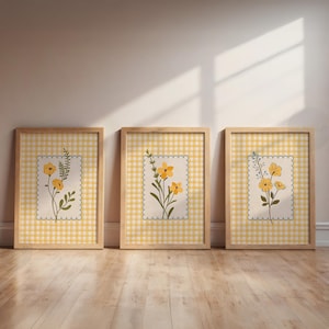 Può includere: Tre stampe artistiche floreali incorniciate. Ogni stampa presenta uno sfondo a quadretti gialli con un quadrato color crema con bordi smerlati. All'interno di ogni quadrato c'è una diversa illustrazione botanica di fiori gialli e foglie verdi. Le cornici sono in legno chiaro.
