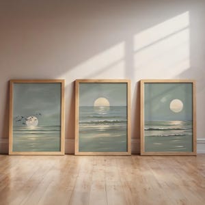 May include: Three framed landscape paintings showing a sunset over the sea. Each artwork features a large, pale yellow sun reflecting on the water, with soft blue and green hues. The frames are made of light-coloured wood, adding a natural touch.