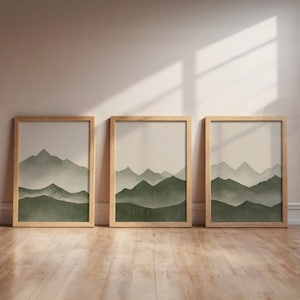 Può includere: Tre stampe artistiche incorniciate raffiguranti un paesaggio montano. Ogni stampa presenta una catena montuosa verde su uno sfondo neutro. Le cornici sono in legno chiaro e le opere d'arte sono esposte su un pavimento in legno.