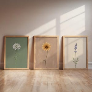 May include: Three framed botanical prints. The prints depict a white flower on a green background, a yellow sunflower on a tan background, and a lavender sprig on a cream background. Each print is in a wooden frame, creating a cohesive set.