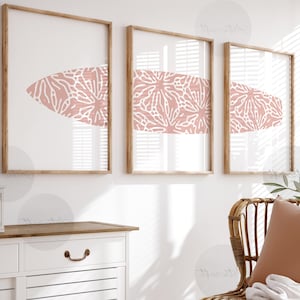 Puede incluir: Tres impresiones artísticas enmarcadas con un diseño en forma de tabla de surf. Cada impresión tiene un marco de madera marrón claro y un fondo blanco. La forma de la tabla de surf es de color rosa coral con un patrón floral blanco.