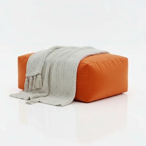 May include: An orange square ottoman with a grey knitted throw blanket draped over it. The blanket has tassels at the ends. The ottoman is a solid colour and appears to be made of a soft material.