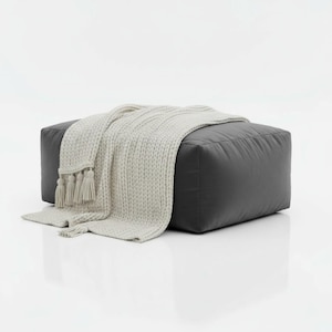 May include: A dark grey, square ottoman with a light grey knitted throw draped over it. The blanket has tassels on one end. The ottoman is a solid colour and appears to be made of leather or a similar material.