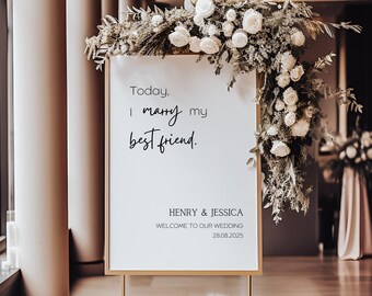 Puede incluir: Un cartel con marco dorado, fondo blanco y texto negro. El texto dice "Today, I marry my best friend. Henry & Jessica. Welcome to our wedding. 28.08.2025". El cartel está decorado con flores blancas y vegetación.