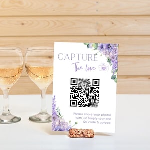 Pode incluir: Um cartaz branco com flores roxas e vegetação no canto superior direito. O cartaz diz "CAPTURE The love" e tem um código QR no centro. O texto abaixo do código QR diz "Compartilhe suas fotos conosco! Basta escanear o código QR e fazer o upload."