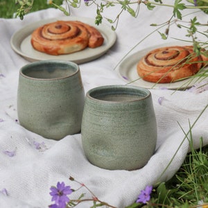 May include: Two green ceramic mugs on a white tablecloth with two cinnamon rolls on white plates. The scene is set outdoors with green grass and purple flowers.