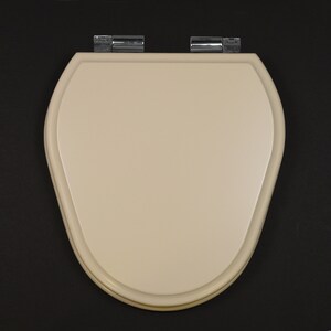 Puede incluir: Un asiento de inodoro beige con forma ovalada redondeada y bisagras cromadas.