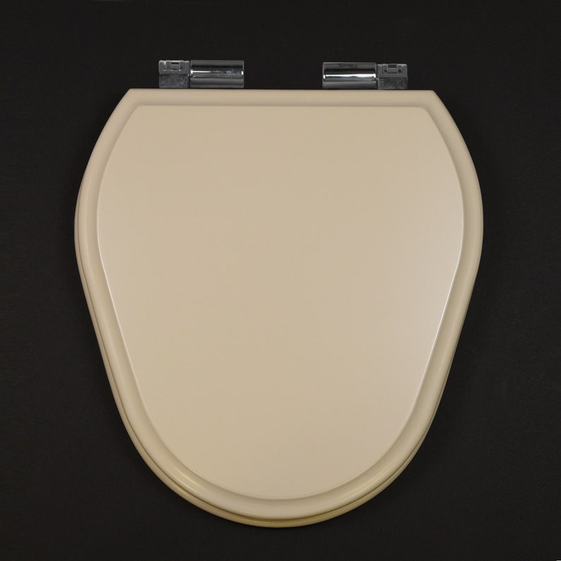 Puede incluir: Un asiento de inodoro beige con forma ovalada redondeada y bisagras cromadas.