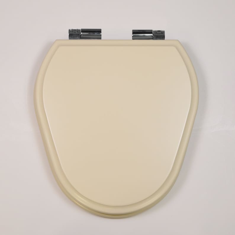 Puede incluir: Un asiento de inodoro de color crema con un borde redondeado y bisagras cromadas.