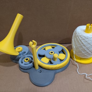 May include: A yellow and gray mechanical yarn winder featuring a central gear assembly, a vertical guide arm, and a conical spindle. A large ball of white yarn sits on a separate yellow gear-shaped base, with a single strand of yarn trailing off.