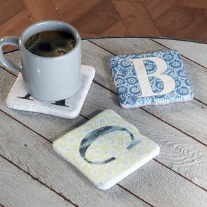 May include: Three square coasters with a white background and a blue, yellow, or white letter on top. The coasters have a floral pattern. A grey mug with coffee is sitting on one of the coasters.