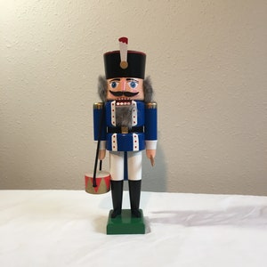 May include: A wooden nutcracker soldier figurine with a blue uniform, white pants, and a red and gold drum. The nutcracker has a black hat with a white plume, a gray beard, and a painted face.