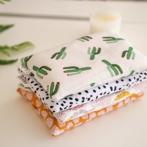 May include: A stack of four fabric heat packs with different patterns. The top pack has a white background with green cactus illustrations. The second pack has a black and white polka dot pattern. The third pack has a pink and white floral pattern. The bottom pack has an orange and yellow geometric pattern.