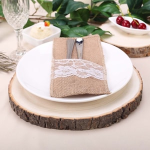 May include: A white plate with a burlap napkin and lace trim holding a fork and knife sits on a wooden slice charger. The charger is on a beige tablecloth.