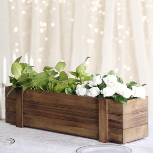 May include: A rustic wooden planter box with a dark brown stain. The planter is filled with green plants and white flowers. The planter is 16 inches long and 6 inches wide.