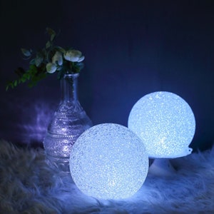 May include: Two white, textured, spherical LED lights with a warm white glow. The lights are on a white fuzzy surface.