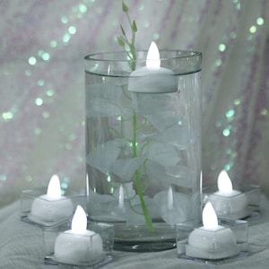 May include: A clear glass vase filled with water and white flower petals, with a single white floating candle in the center. Four additional white floating candles are arranged on clear plastic bases around the vase.