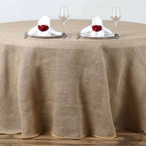 May include: A round, brown burlap tablecloth with a natural, rustic look. Two place settings are set on the tablecloth with white napkins and red rose napkin rings.
