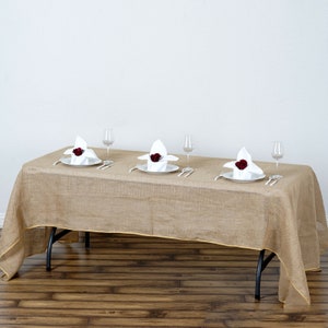 May include: A rectangular tan burlap tablecloth with a gold trim, set for a formal dinner. Each place setting includes a white plate, folded napkin with a red rose, silverware, and a wine glass. The table is on a wooden floor.
