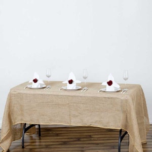 May include: A rectangular brown burlap tablecloth with a gold trim, set on a wooden table with three place settings. Each place setting includes a white napkin with a red rose, a white plate, a fork, a knife, and a wine glass.