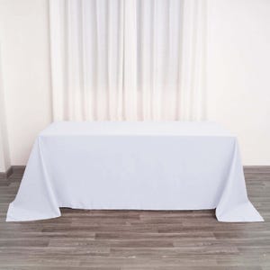 May include: A white rectangular tablecloth draped over a table. The fabric is smooth and covers the table completely, with the edges hanging down. The background features white curtains and a wooden floor.