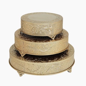 May include: A set of three gold-colored round cake stands with an embossed floral design. The stands are stacked on top of each other, with the largest stand at the bottom and the smallest stand at the top.