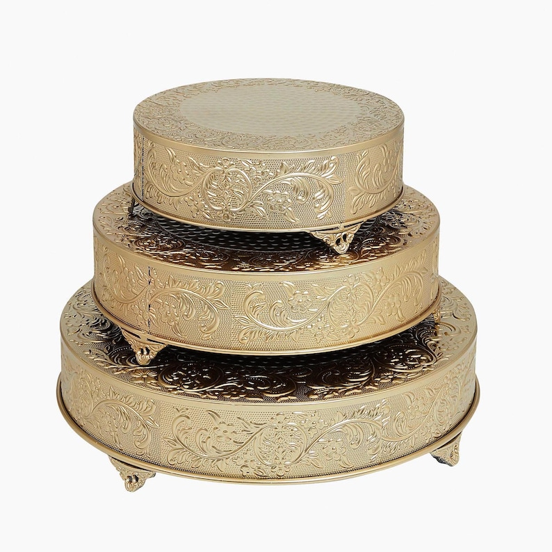 May include: A set of three gold-colored round cake stands with an embossed floral design. The stands are stacked on top of each other, with the largest stand at the bottom and the smallest stand at the top.