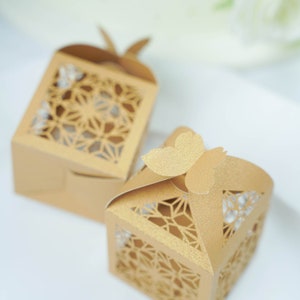 May include: Two gold-colored favor boxes with intricate geometric cutouts. One box is open, and the other is closed with a butterfly-shaped closure. The boxes are made of a textured paper material, perfect for small gifts or treats.