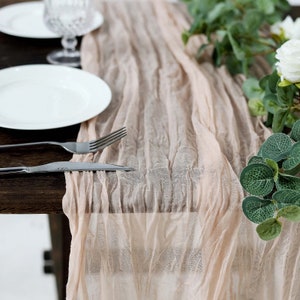 May include: A light pink sheer table runner with a crinkled texture. The runner is laid on a wooden table with a white plate and silverware.