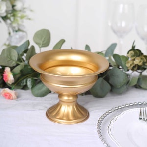 May include: A gold-colored metal pedestal bowl with a wide, shallow top and a fluted base. The bowl is designed for decorative purposes, possibly for centerpieces or floral arrangements. The background includes greenery and a white tablecloth.