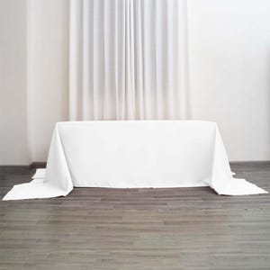 May include: A rectangular table covered with a solid white tablecloth that drapes over the sides and ends, reaching down to the floor. The fabric is smooth and opaque, providing full coverage for the table surface and legs in a clean, neutral style.
