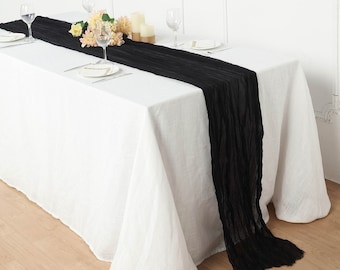 May include: A black table runner with a crinkled texture is placed on a white tablecloth. The runner is long and drapes over the edge of the table.