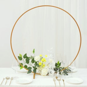 May include: A gold metal hoop arch with white and yellow flowers and greenery. The arch is standing on a white table with white plates and silverware.