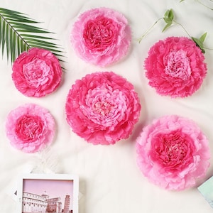 May include: Six pink paper flowers of varying sizes. The flowers are arranged on a white surface.