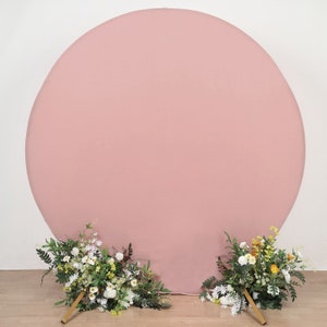 May include: A large, round, blush pink backdrop for photography. The backdrop is made of fabric and is perfect for creating a soft, romantic look. Two bouquets of white and yellow flowers are placed in front of the backdrop.